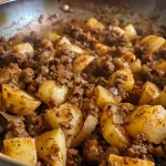 A close-up view of a skillet filled with ground beef and cubed potatoes, garnished with onions and garlic.