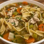 A close-up view of a bowl of steaming homestyle chicken noodle soup with visible noodles and vegetables.