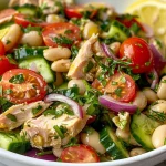 A colorful bowl of white bean and tuna salad with lemon slices, cherry tomatoes, and herbs.