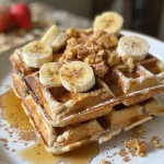 A stack of golden-brown flourless banana oat waffles topped with fresh fruit and maple syrup.