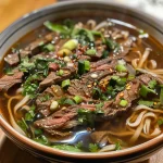 A close-up side view of a steaming bowl of spicy beef noodle soup with vibrant ingredients.