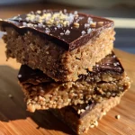 ['Close-up of gluten-free, dairy-free protein bars drizzled with melted dark chocolate.', 'Side view of homemade protein bars showcasing chunks of chocolate and a sprinkle of sea salt.', 'Delicious protein bars with a glossy chocolate coating and visible seeds on top.', 'An appetizing arrangement of protein bars with a rich chocolate finish and nutty texture.']