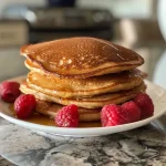 ['Close-up of golden-brown keto pancakes stacked on a plate.', 'Side view of fluffy keto pancakes drizzled with syrup.', 'Tasty keto pancakes on a white plate with a fork beside them.', 'Delicious low-carb pancakes topped with blueberries and syrup.']