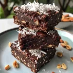 ['Close-up view of a stack of No Bake Coconut Brownies with chocolate pieces on top.', 'Side view of No Bake Coconut Brownies cut into squares, showcasing the coconut texture.', 'Juicy No Bake Coconut Brownies arranged on a white plate, garnished with chocolate chips.', 'A delicious slice of No Bake Coconut Brownies with visible layers of coconut and chocolate.']