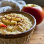 ['Close-up of a creamy apple rice pudding with apple slices and a sprinkle of cinnamon.', 'Angular view of a bowl filled with apple rice pudding topped with grated apple and a drizzle of maple syrup.', 'Side view of apple rice pudding showing its texture and ingredients, including red apple slices.', 'Detailed close-up of a serving of apple rice pudding with cardamom and coconut oil visible.']