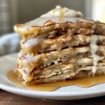 ['Close-up view of golden brown cinnamon roll pancakes stacked with drizzled maple syrup.', 'Side view of fluffy cinnamon roll pancakes topped with cream cheese frosting and sprinkled cinnamon.', 'Juicy cinnamon roll pancakes on a plate, featuring a glossy maple syrup glaze and a side of fresh berries.', 'Appetizing cinnamon roll pancakes with a creamy topping, surrounded by a warm kitchen setting.']