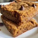 ['Side view of a decadent Pumpkin Spice Blondie, showcasing its moist texture.', 'Close-up of golden-brown Pumpkin Spice Blondies topped with chocolate chips.', 'Delicious Pumpkin Spice Blondies in a baking dish, highlighting their chewy consistency.', 'A slice of Pumpkin Spice Blondie with a rich color and chocolate chip garnish.']