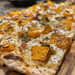 ['Close-up view of a slice of roasted butternut squash pizza topped with crispy sage.', 'Side profile of a delicious roasted butternut squash pizza, showcasing its toppings.', 'Juicy roasted butternut squash pizza with fresh ricotta and sage leaves prominently displayed.', 'A mouthwatering slice of butternut squash pizza highlighted by golden roasted squash and herbs.']