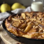 ['Close-up of a spiced pear and walnut Dutch baby pancake garnished with powdered sugar.', 'Side view of a fluffy Dutch baby topped with sliced pears and walnuts on a plate.', 'A delicious Spiced Pear & Walnut Dutch Baby served with lemon wedges and crème fraîche.', 'A golden-brown Dutch baby pancake decorated with cinnamon and brown sugar on a rustic table.']